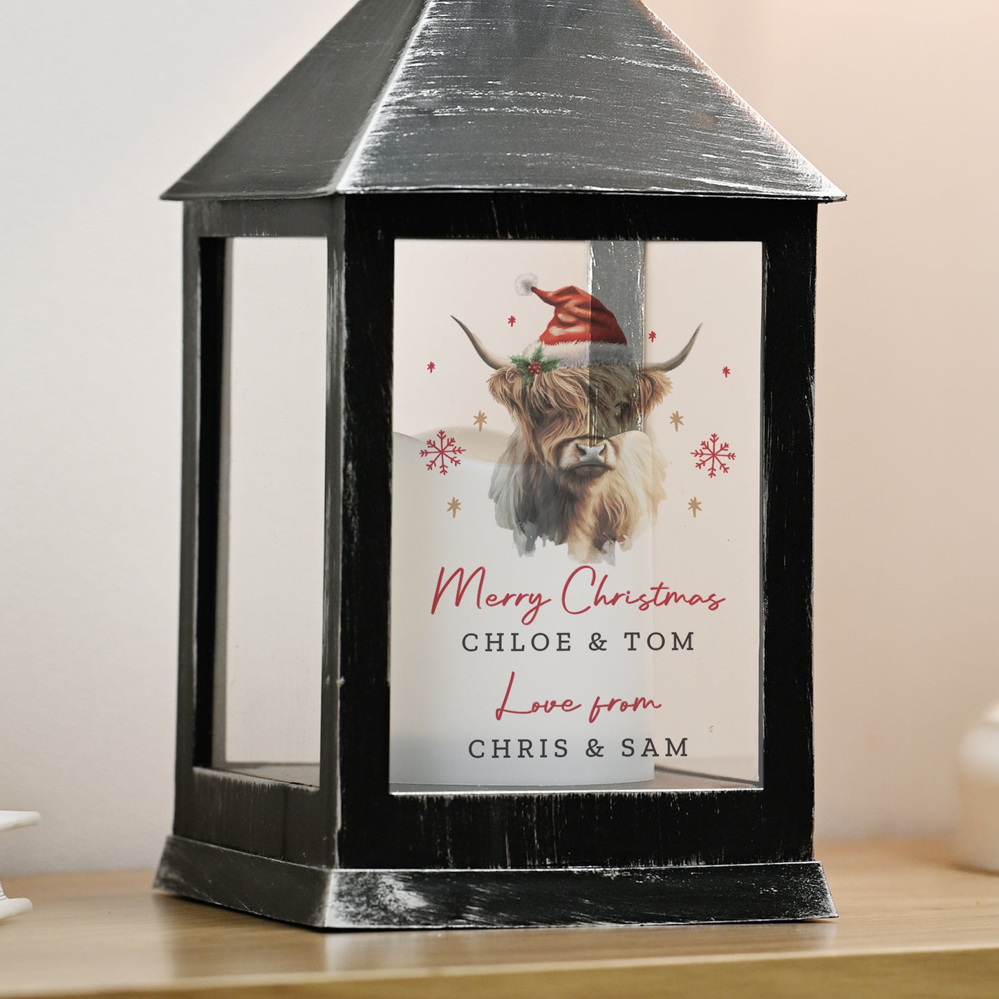 Black lantern with a Christmas card featuring a highland cow wearing a Santa hat on a wooden surface.