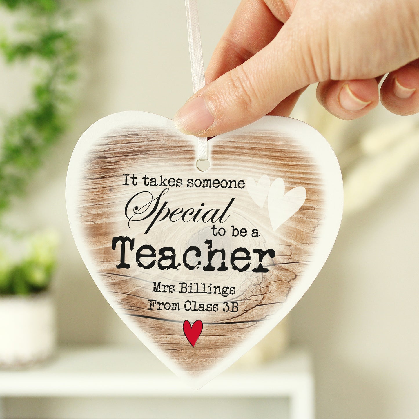 Heart-shaped wooden ornament with printed text, held by a hand against a blurred indoor background.