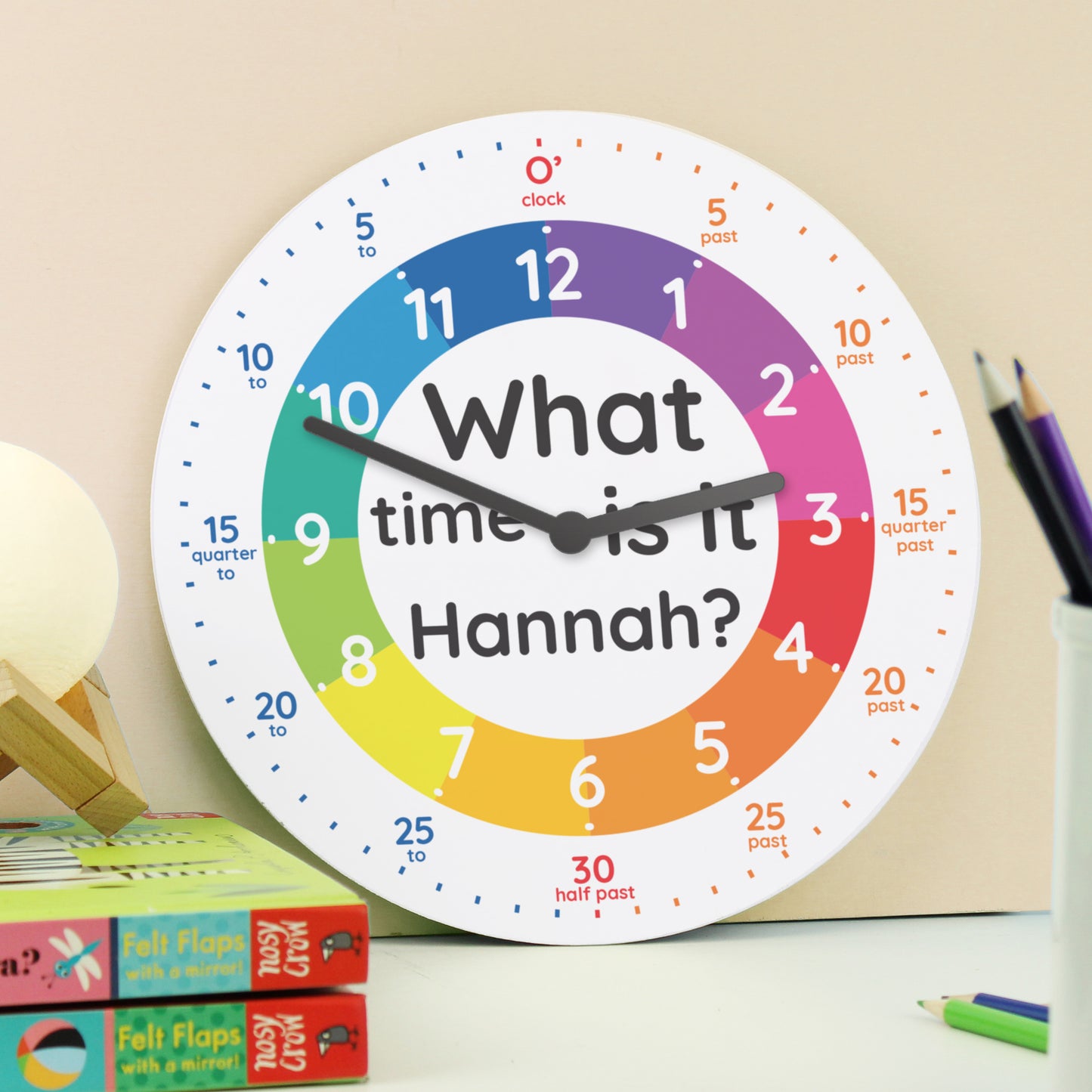 Colourful clock with 'What time is it Hannah?' text on a desk with educational materials.