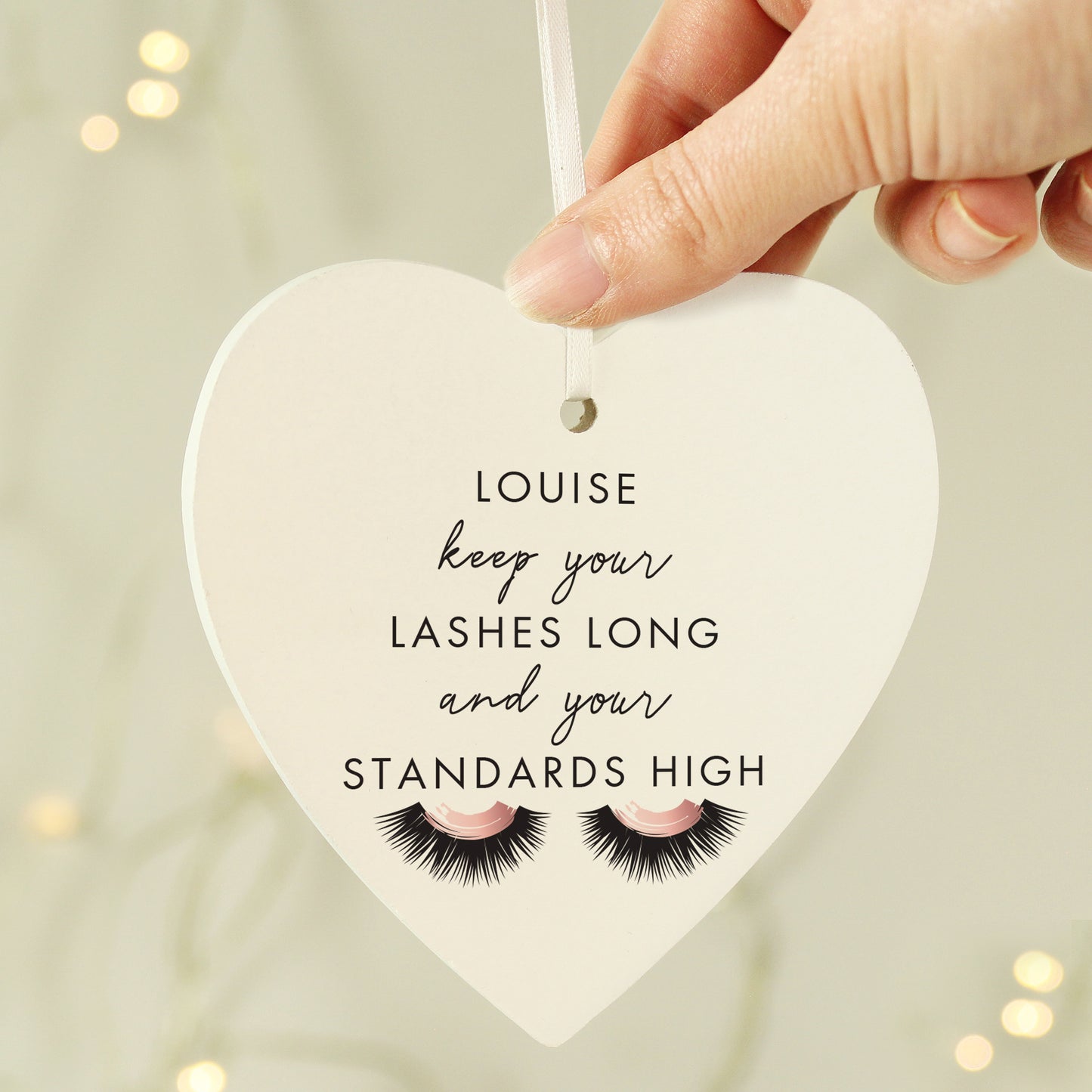 Heart-shaped sign with motivational quote and false eyelashes, held by a hand against a blurred background.