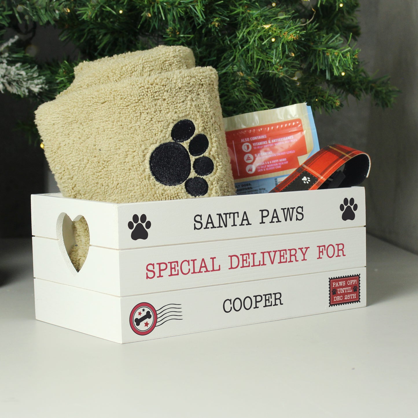White wooden crate labeled 'Santa Paws' with pet supplies under a Christmas tree.
