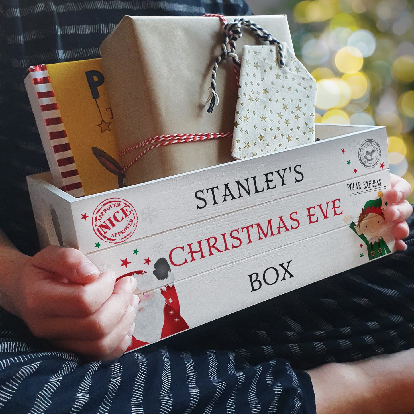 Person holding a 'Stanley's Christmas Eve Box' filled with gifts against a blurred festive background.