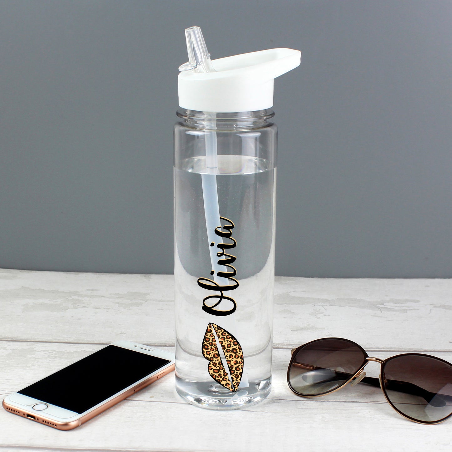 Clear water bottle with 'Olivia' and leaf design, smartphone, and sunglasses on a light surface.