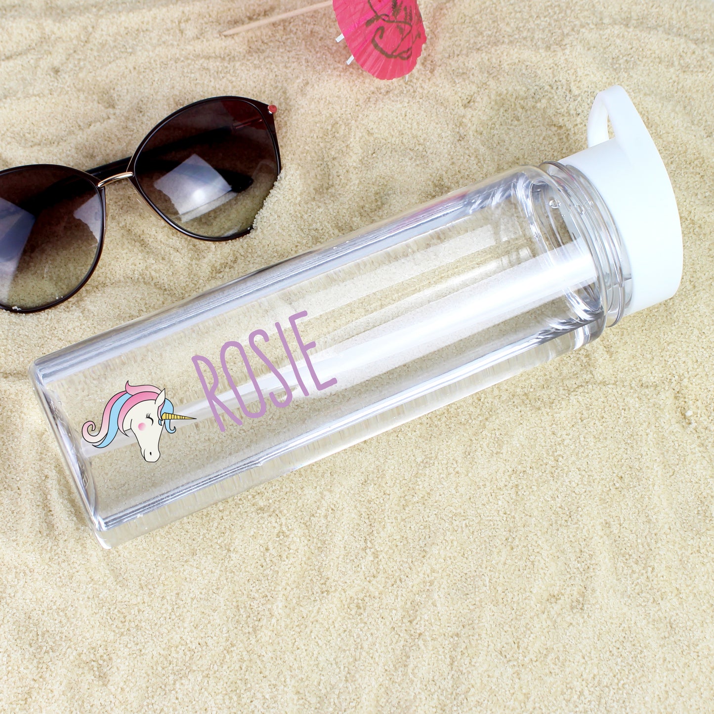Clear water bottle with unicorn design and 'Rosie' text on sandy surface with sunglasses and pink item.