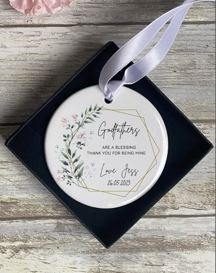 Close-up of a round ornament reading “Godfathers are a blessing, thank you for being mine. Love Jess 26.05.2023” inside a navy gift box with floral graphics and ribbon.