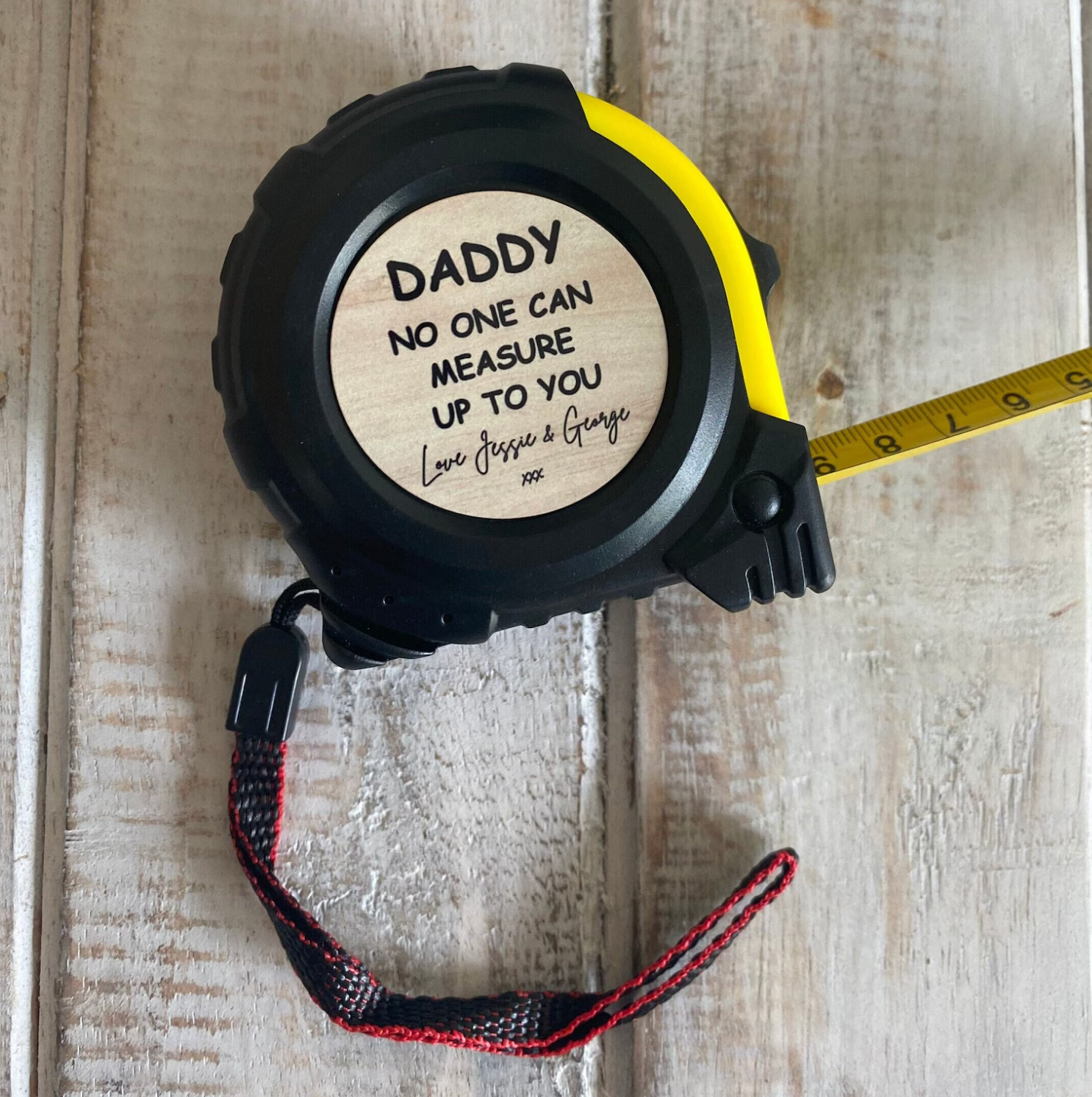 A black and yellow tape measure with a wooden label that reads, “DADDY – No one can measure up to you. Love Jessie & George xxx,” resting on a rustic wooden surface with the measuring tape partially extended and a red and black wrist strap attached.