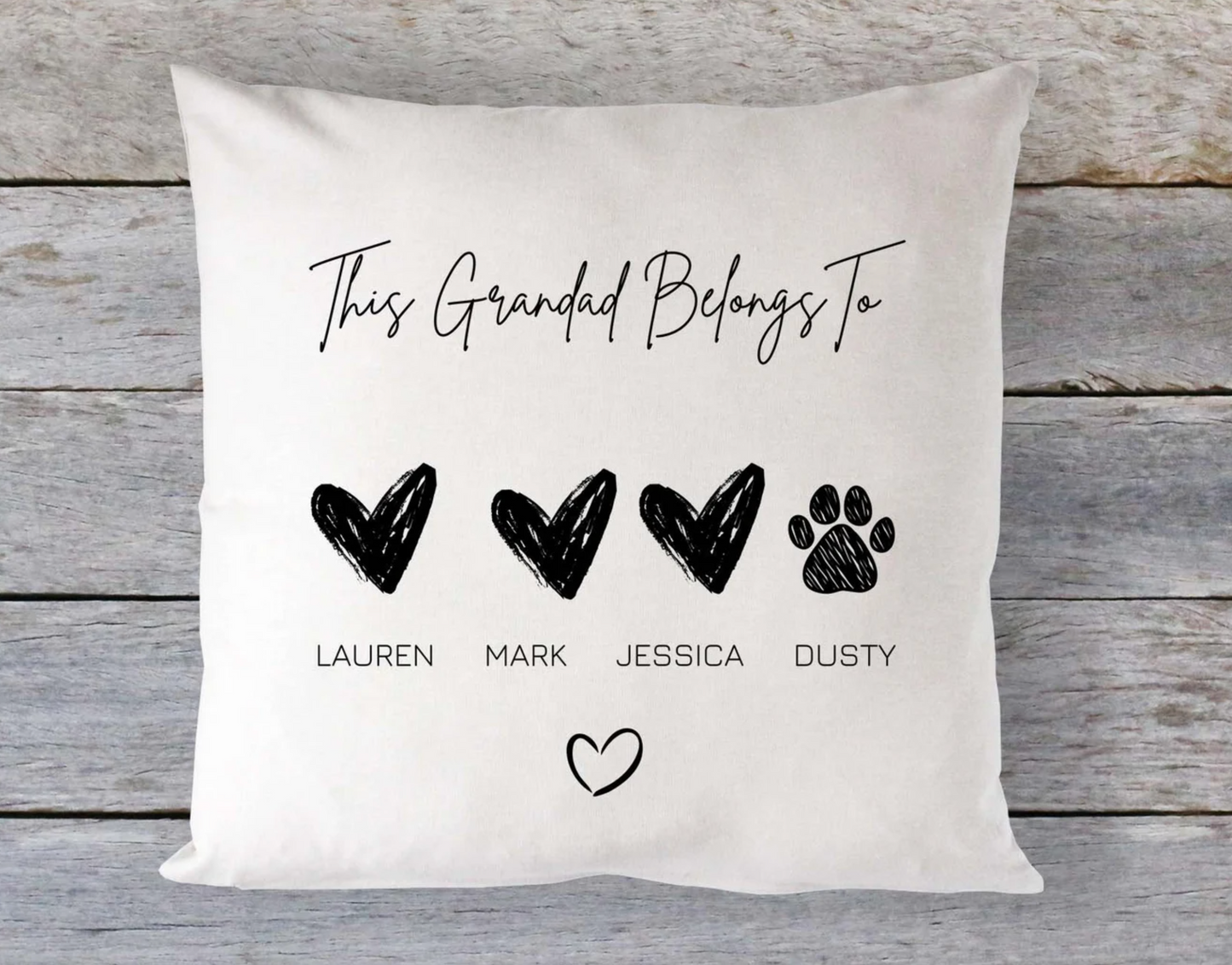 Square cushion with light beige fabric featuring the phrase “This Grandad Belongs To” in a handwritten-style black font at the top. Below are three large black sketched hearts labeled “LAUREN,” “MARK,” and “JESSICA,” and a black paw print labeled “DUSTY.” A smaller heart icon is centered at the bottom of the design. The cushion is placed against a rustic wooden background.