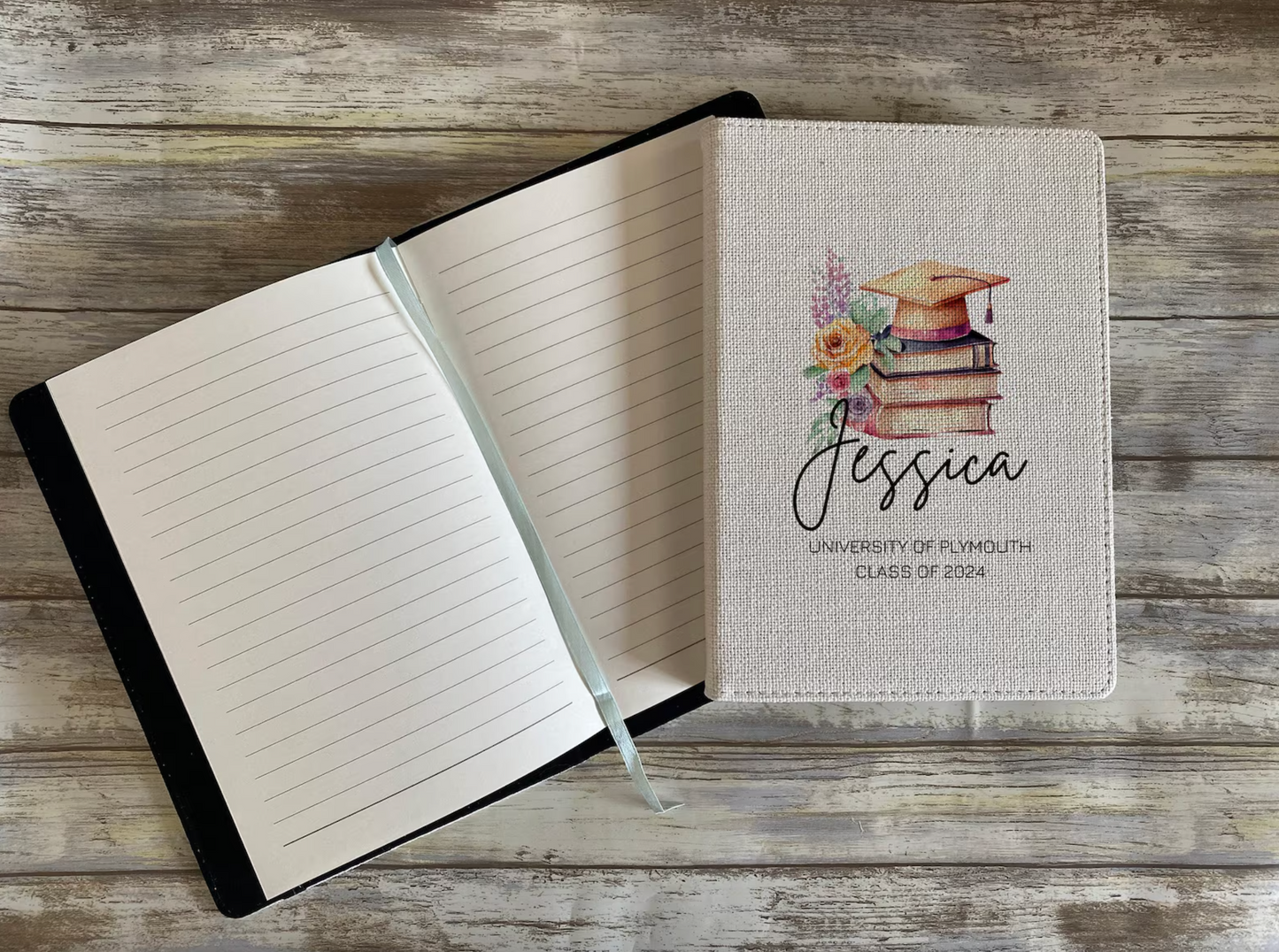 A graduation-themed notebook displayed on a rustic wooden surface. The closed cover of the right notebook features a printed illustration of a stack of vintage books topped with a golden graduation cap and adorned with flowers. The name “Jessica” is written in an elegant script font, with “University of Plymouth, Class of 2024” printed below in uppercase. To the left, an open notebook with lined pages and a pale blue ribbon bookmark is shown.