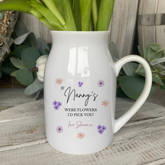 A white ceramic jug filled with fresh green tulips, featuring a sentimental printed message that reads: “If Nanny’s were flowers, I’d pick you! Love Shauna xx” in elegant script. The jug is decorated with scattered pink, purple, and lavender floral accents and is placed on a rustic wooden surface surrounded by soft greenery.