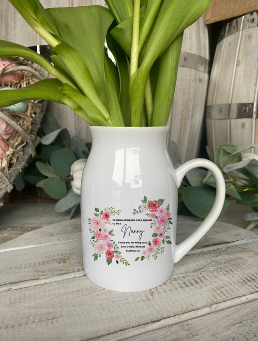 A white ceramic jug filled with fresh green tulip stems and buds, featuring a floral heart-shaped wreath design on the front. Inside the wreath, the text reads: "It takes someone very special to be a Nanny. Thank you for being ours. Love Jessie, Michael & Arthur xx." The jug is placed on a rustic wooden surface, surrounded by soft decor and greenery.