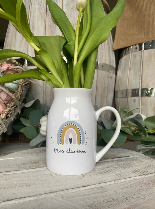 A white ceramic jug used as a vase, filled with green tulip stems and buds. The jug is personalized with the name "Mrs Clarkson" written in a cursive font below a pastel rainbow design decorated with stars and dots. The jug is placed on a rustic wooden surface with a heart-shaped wicker decoration and artificial greenery in the background.