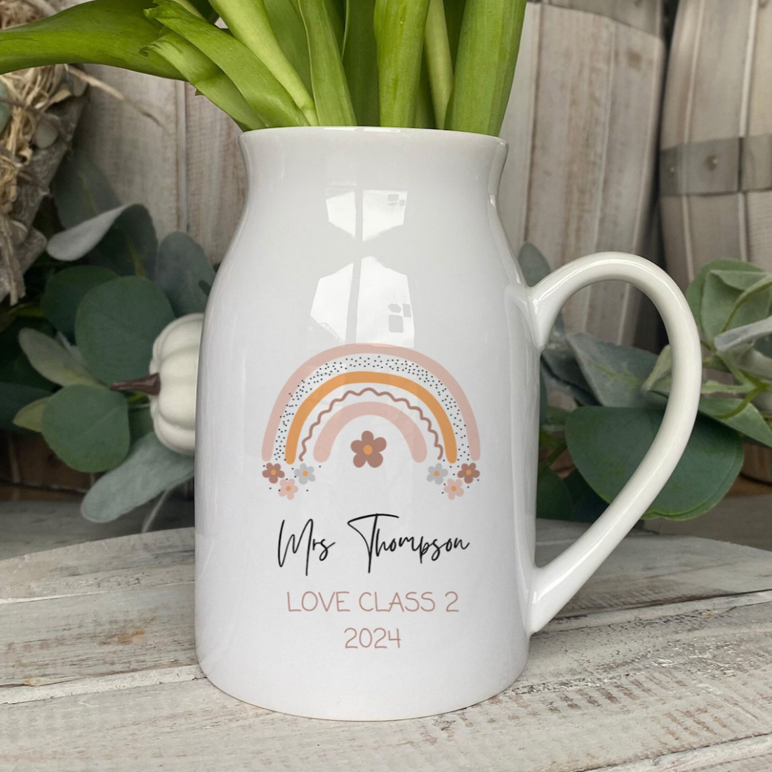 A white ceramic vase filled with tulips, personalized with the name “Mrs Thompson” in black script. Below the name, text reads “LOVE CLASS 2 2024” in peach. The vase is decorated with a pastel rainbow and floral design. It is placed on a rustic wooden surface with greenery and soft-focus background elements including a wooden barrel and a heart-shaped wreath.