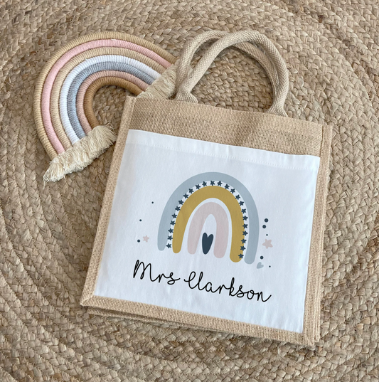 Personalised jute tote bag laid on a woven mat, featuring a pastel rainbow in yellow, pink, and grey shades. Underneath the design is the name "Mrs Clarkson" in a fun, handwritten font. A fabric rainbow decoration is placed beside the bag.