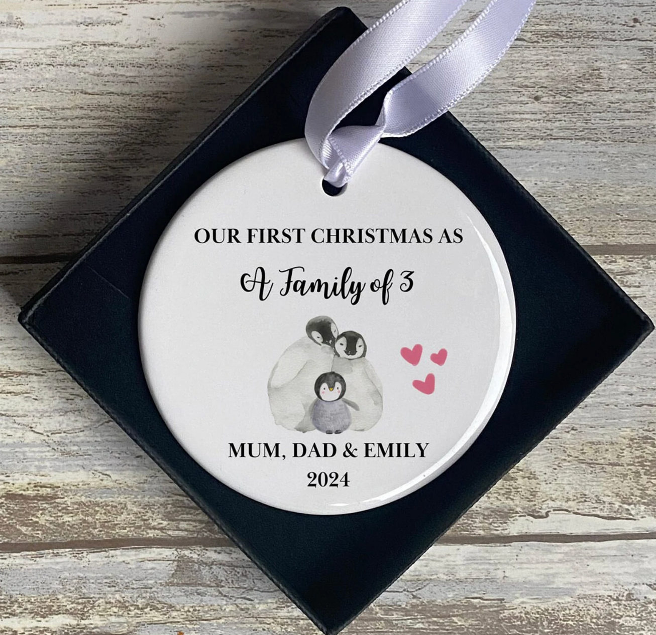 Personalised ceramic Christmas ornament in gift box, with illustration of a penguin family of three and pink hearts. Custom text reads: “Our First Christmas As A Family of 3 – Mum, Dad & Emily – 2024.” Ideal keepsake for new parents celebrating baby’s first Christmas.