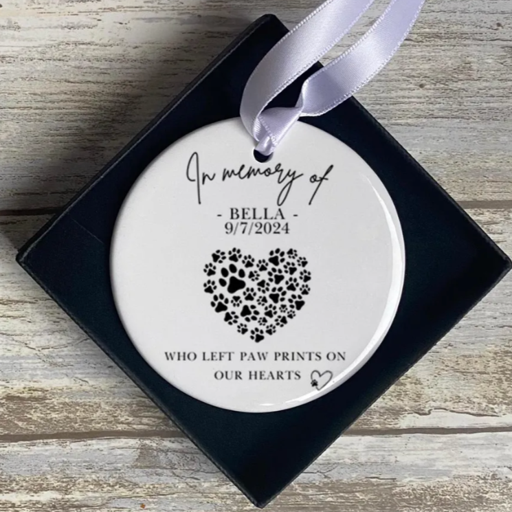 A close-up of a round white ceramic ornament with a satin ribbon, displayed in a black gift box. The ornament reads “In memory of Bella - 9/7/2024” above a heart shape made from various black paw prints. Below the heart is the phrase “Who left paw prints on our hearts” with a small heart illustration.