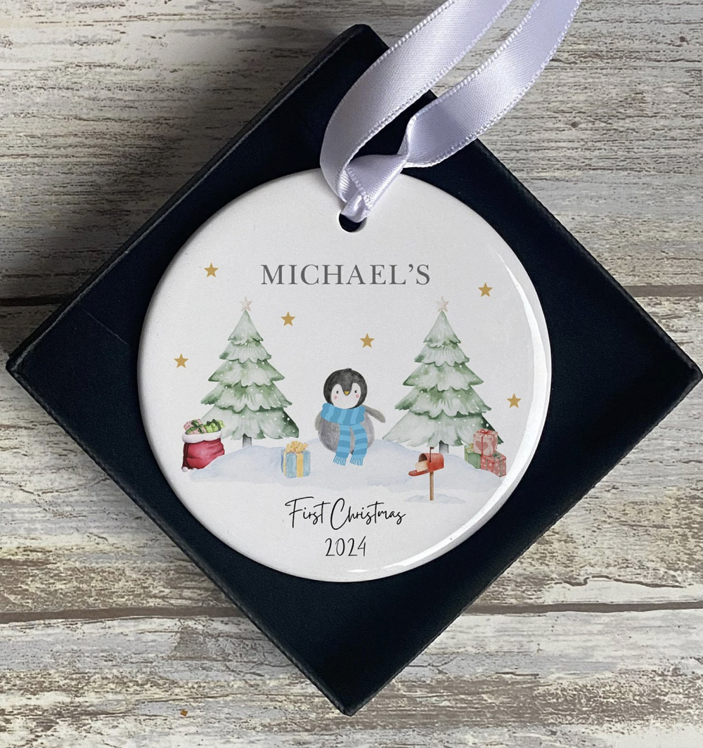 Custom Christmas tree ornament with a winter scene, personalised with “Michael.” Includes a sweet penguin in blue, gift-wrapped presents, and snow-covered trees. A first Christmas 2024 ceramic bauble ideal for baby boys or toddlers, packaged in a premium presentation box.