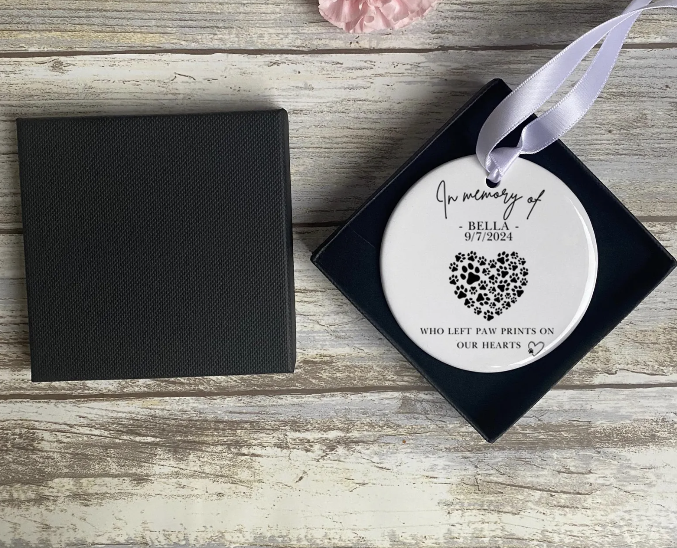 Personalised pet memorial ornament featuring the name “Bella” and the date “9/7/2024,” with the phrase “In memory of” above and “Who left paw prints on our hearts” below a heart-shaped paw print design. The ceramic decoration hangs from a white ribbon and is displayed in a black gift box.