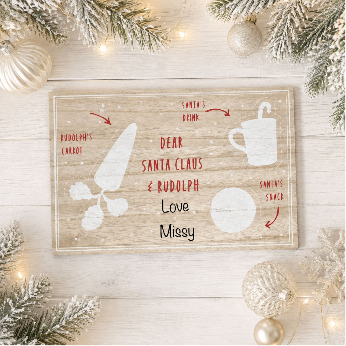 A rectangular rustic wooden santa treat board with a Christmas-themed letter design, styled in a flat-lay on a white wood background with holiday decorations including pine branches, ornaments, and fairy lights.