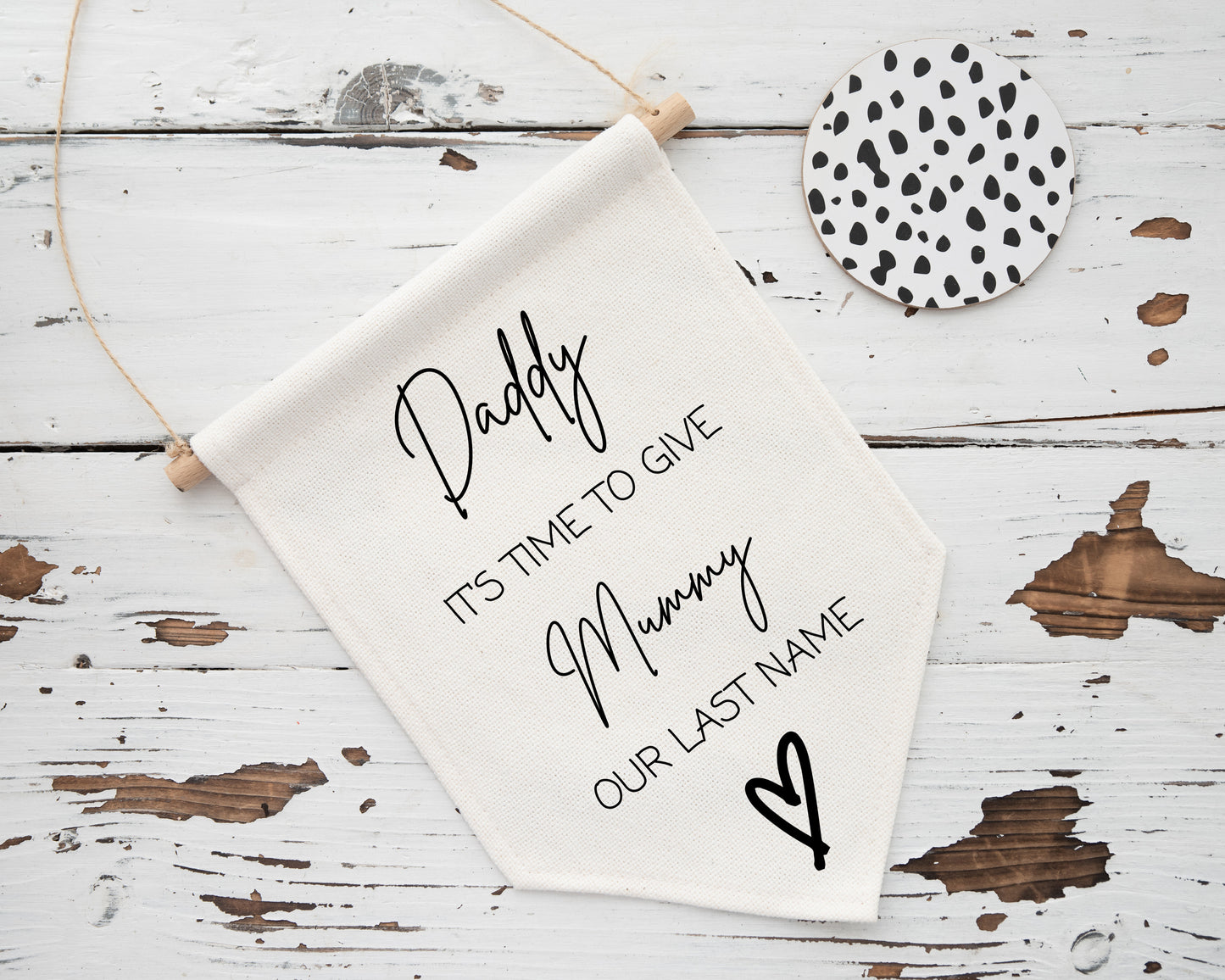 Cream fabric banner with black text that reads: “Daddy, it’s time to give Mummy our last name” in a mix of elegant cursive and block fonts. A black heart illustration is placed in the bottom corner. The banner is laid on a rustic white wooden background with a spotted round coaster nearby.