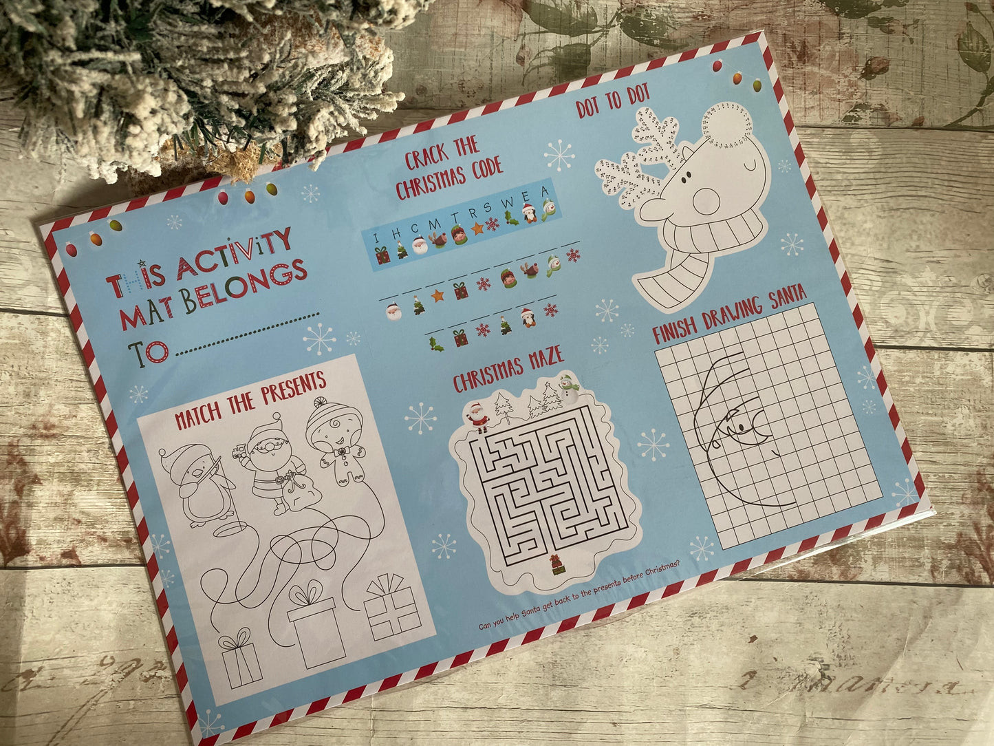 A colourful Christmas-themed children's activity mat with various fun games printed on a light blue background. The mat includes sections like “This activity mat belongs to,” “Crack the Christmas Code,” “Dot to Dot,” “Finish Drawing Santa,” “Christmas Maze,” and “Match the Presents.” Surrounded by candy cane stripe borders, the mat is filled with festive illustrations such as Santa, elves, a reindeer, and snowflakes. A perfect holiday entertainment item for young children.