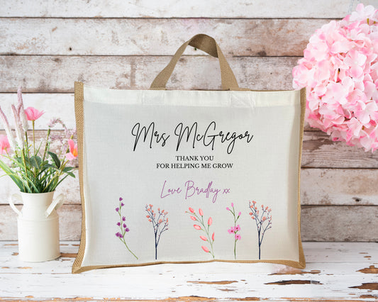 Personalised jute teacher gift bag with beige canvas front, featuring the text “Mrs McGregor – Thank you for helping me grow – Love Bradley xx” in elegant black and purple script fonts. The design includes hand-drawn floral illustrations in pink, purple, and coral tones along the bottom. The bag is styled against a rustic wooden background with pink flowers and a white jug of wildflowers beside it.