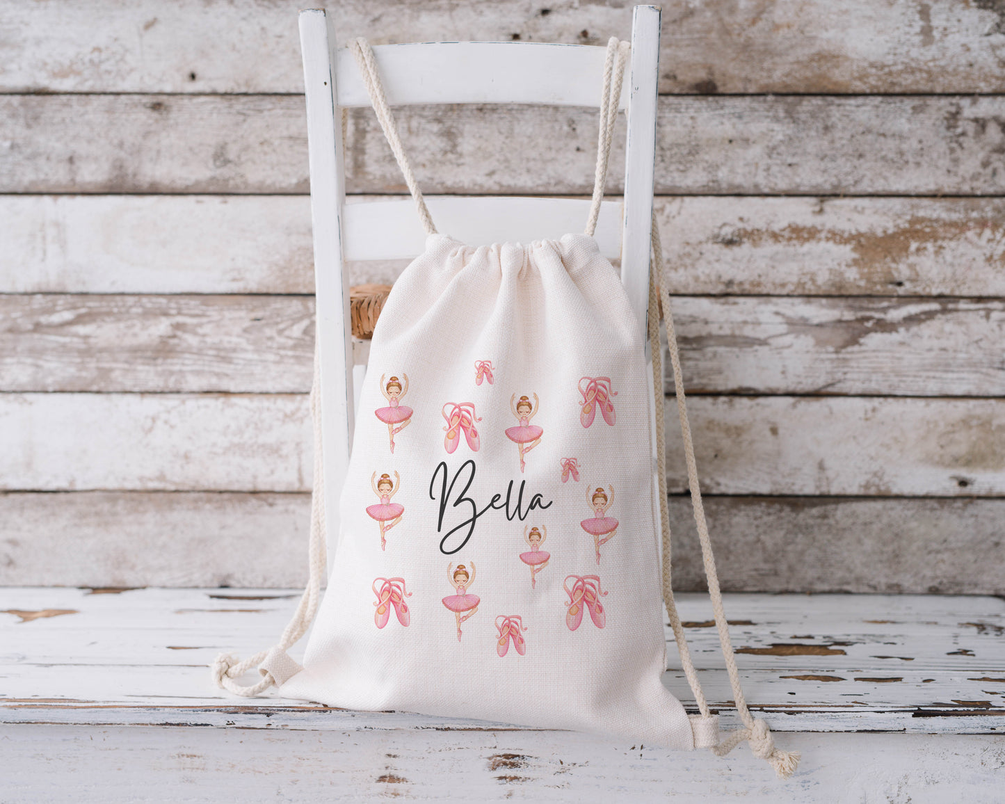 Children’s personalised drawstring ballet bag shown hanging on a white wooden chair, featuring a pink ballerina and ballet slipper pattern with the name “Bella” written in a stylish black script font. Perfect for dance classes or as a gift for young ballerinas.