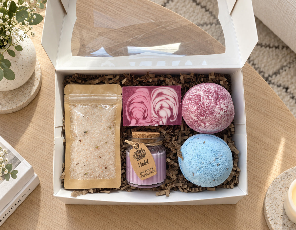 A curated bath gift set in a white box with a clear window lid, arranged on a wooden table.