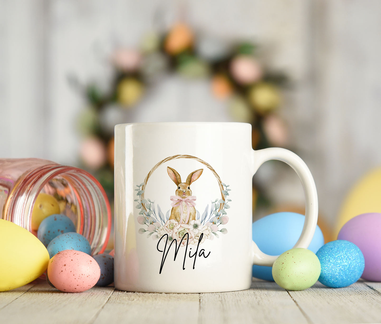 Personalized Easter mug featuring a watercolor bunny with a pink bow inside a floral hoop wreath, custom name "Mila" below the design. Surrounded by colorful Easter eggs and a rustic background.