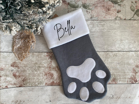 Custom grey pet stocking shaped like a paw, featuring a white cuff with the name “Bella” in script. A cute and festive Christmas accessory for dogs or cats to hold treats and toys.