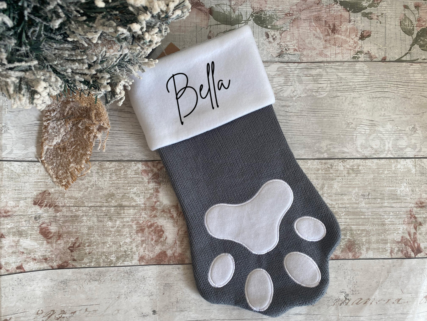 Personalised pet Christmas stocking in a grey paw shape with white felt paw print details. The white cuff is embroidered with the name “Bella,” perfect for including furry family members in festive celebrations.