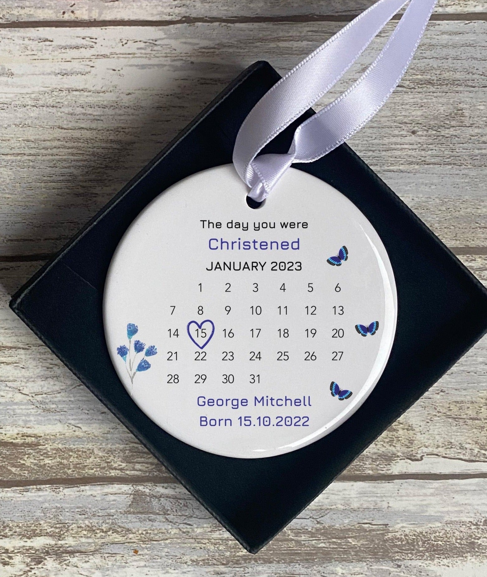 Personalised ‘The Day You Were Christened’ ornament featuring a calendar for January 2023, with the date 15th marked by a heart. The ornament has a white background, blue text, and blue butterfly and floral details. It is presented in a black gift box, making it a unique, personalised gift for a christening.