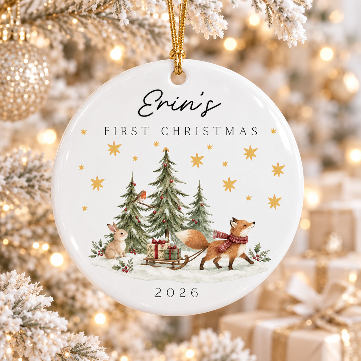 A personalized round ceramic Christmas ornament hanging from a gold string, featuring the text 'Erin's FIRST CHRISTMAS 2026' and an illustration of a fox pulling a sleigh with gift boxes near decorated trees. The ornament is shown hanging in a softly blurred festive tree setting with warm lights and wrapped gifts.