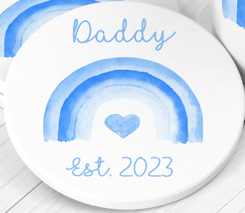 Round white coaster with blue rainbow design and 'Daddy Est. 2023' text on a light wooden surface.