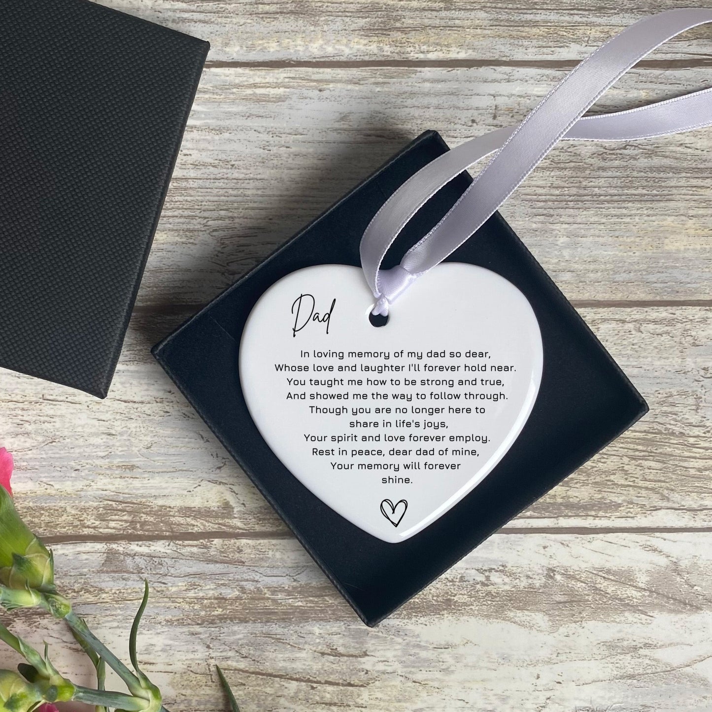 A heart-shaped, white ceramic ornament placed in an open black gift box. The ornament has a printed poem about remembrance for a father, starting with the word "Dad" in cursive. A grey ribbon is tied to the ornament for hanging, and the gift box rests on a rustic wooden surface next to a few pink flowers.