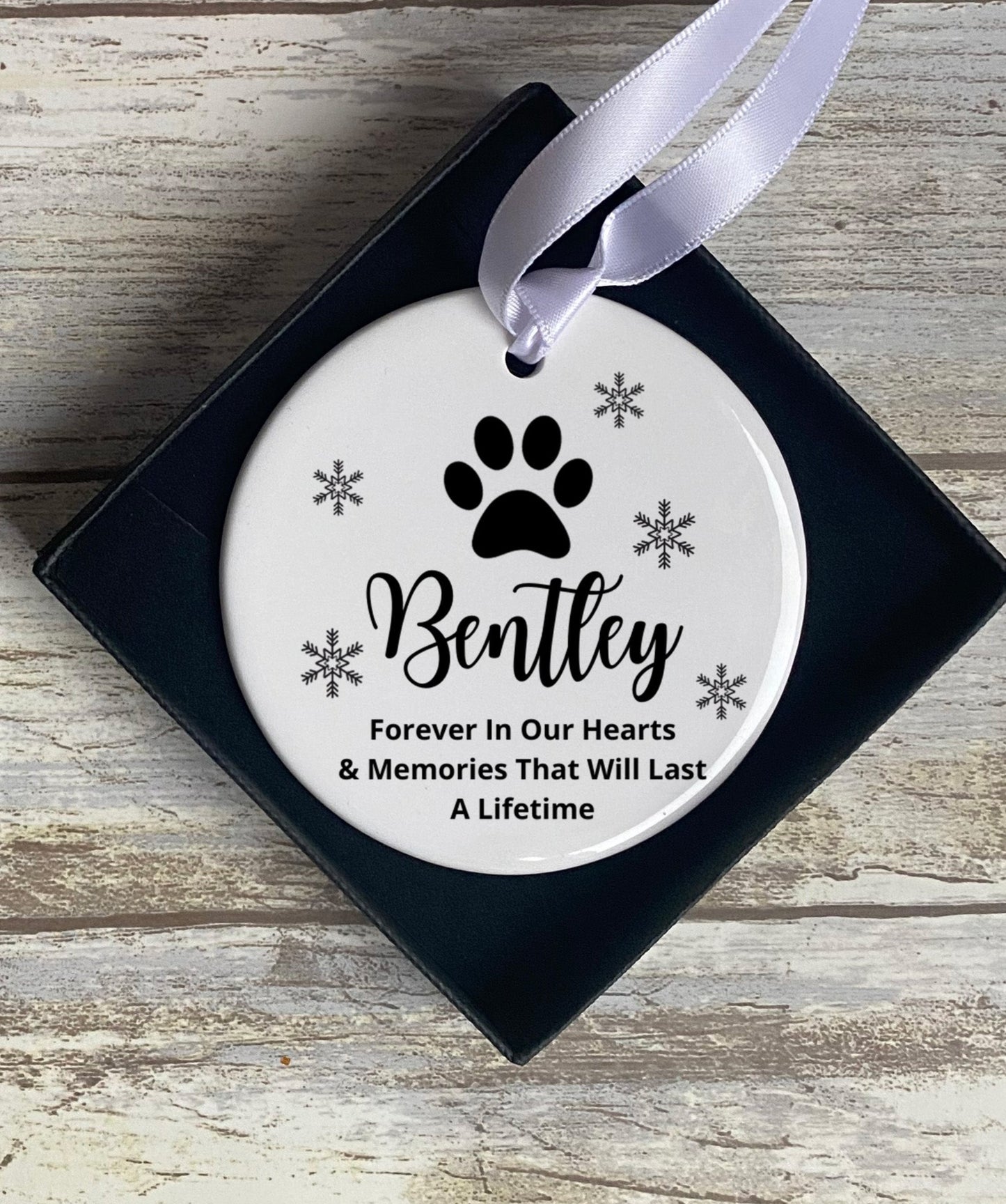 A round, white ceramic Christmas ornament with black text and design, featuring a paw print and snowflake motif, with a personalized message for a pet.