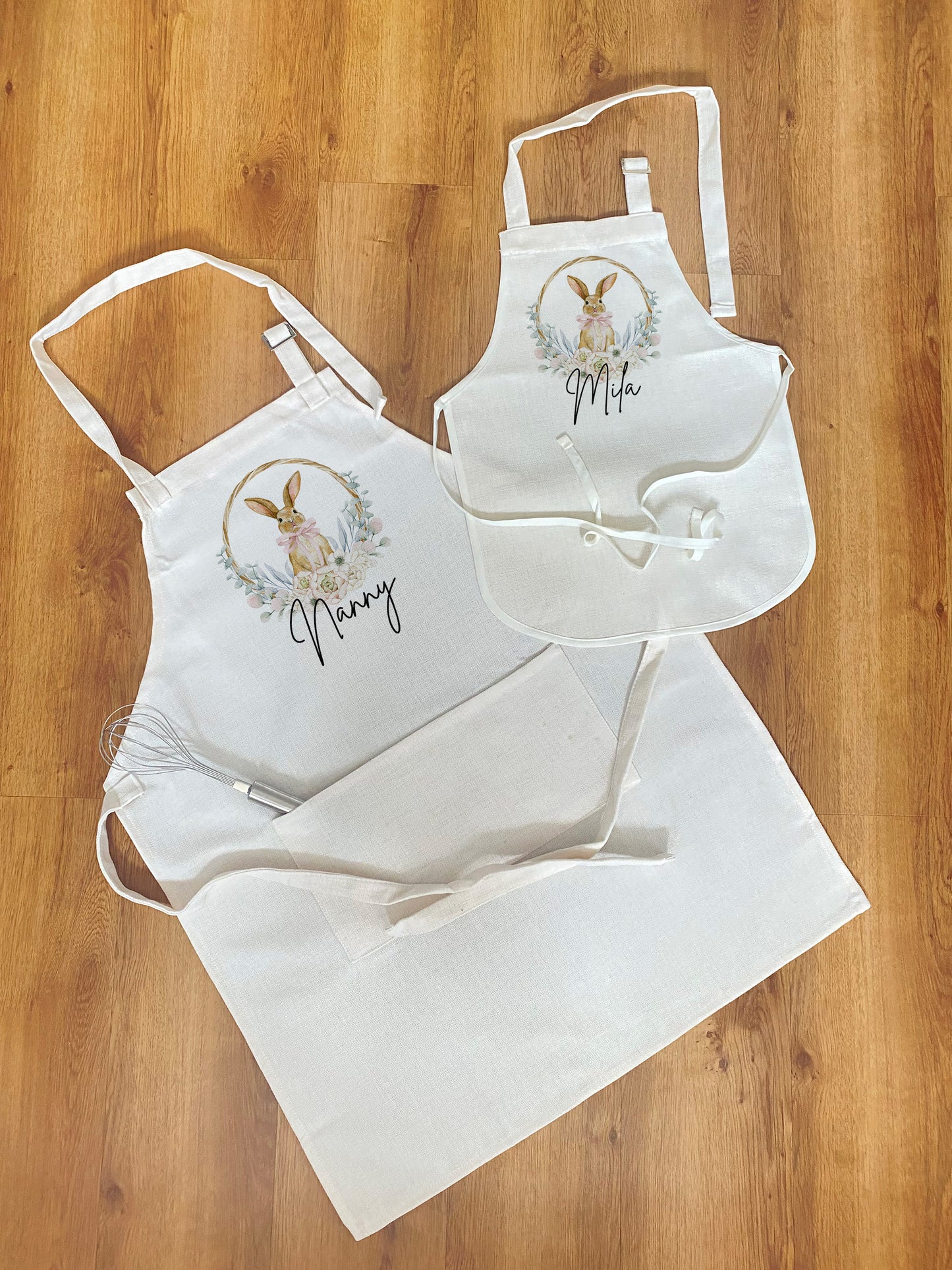 Flat lay of two matching white aprons on a wooden floor — one adult-sized with the name “Nanny” and one child-sized with the name “Mila.” Both aprons feature a cute watercolor bunny inside a floral wreath. A metal whisk is placed in the pocket of the adult apron.
