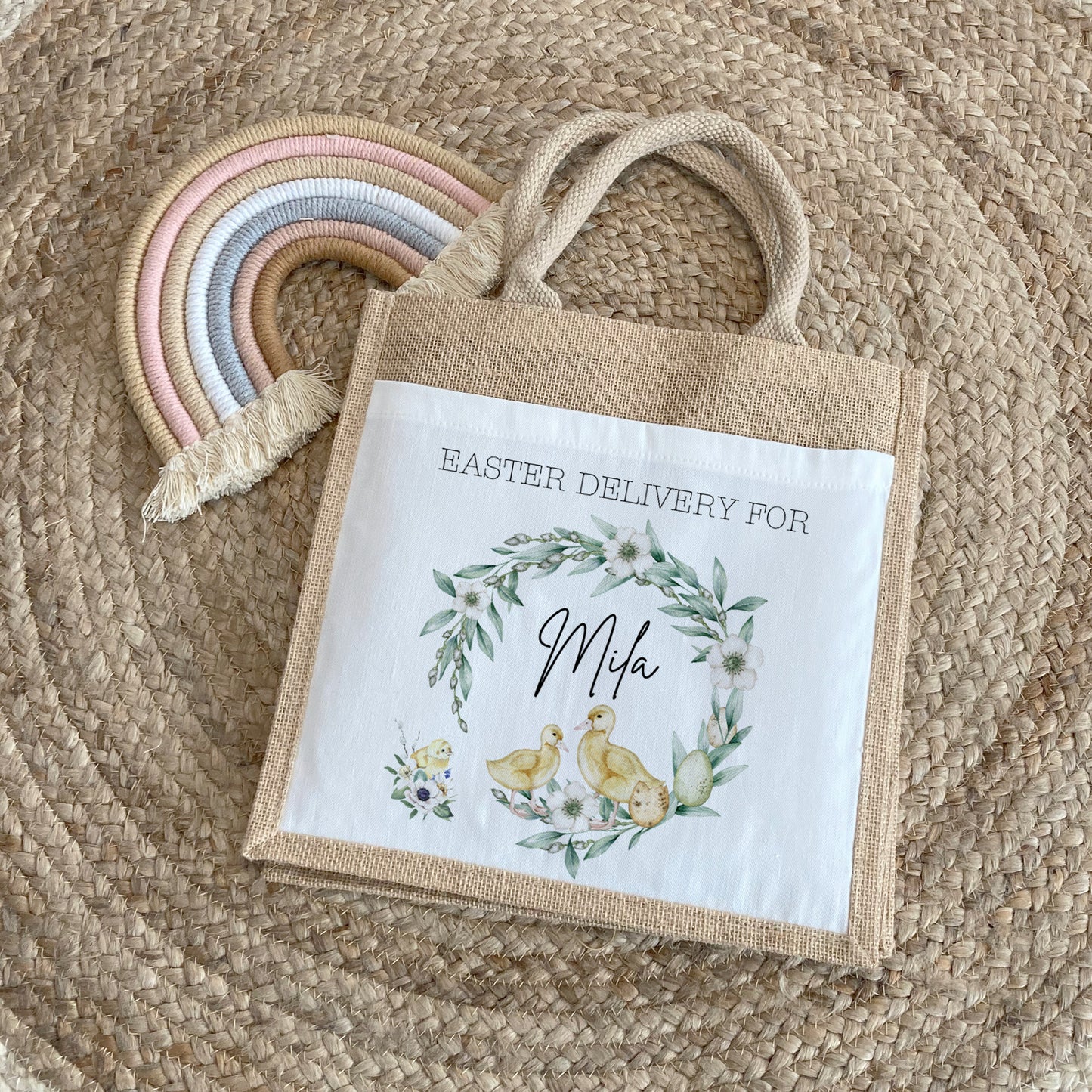 Personalized jute Easter tote bag with the name “Mila” printed on a white front panel. The design features a wreath of green foliage and white flowers surrounding three yellow chicks and speckled Easter eggs. Above the wreath, it says “EASTER DELIVERY FOR.” The bag is placed on a woven jute rug next to a pastel-colored rainbow decoration.
