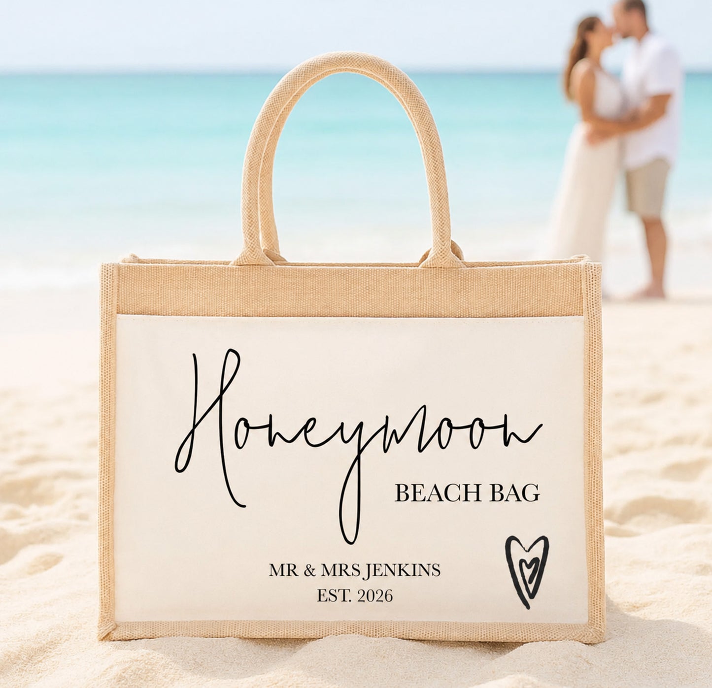 A personalized jute beach tote bag with a cream insert and black printed text reading 'Honeymoon Beach Bag' and 'Mr & Mrs Jenkins Est. 2026', set on a sandy beach with a blurred couple and ocean in the background.