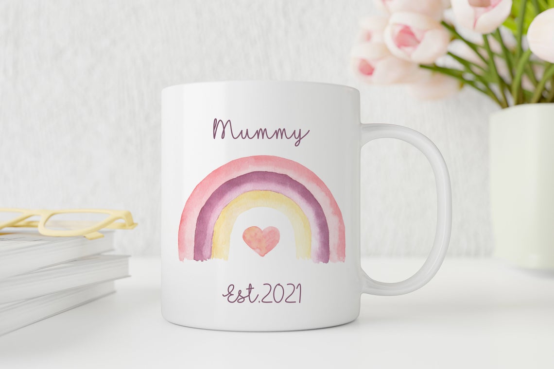 White mug with a rainbow and heart design, text 'Mummy est 2021', on a light background.