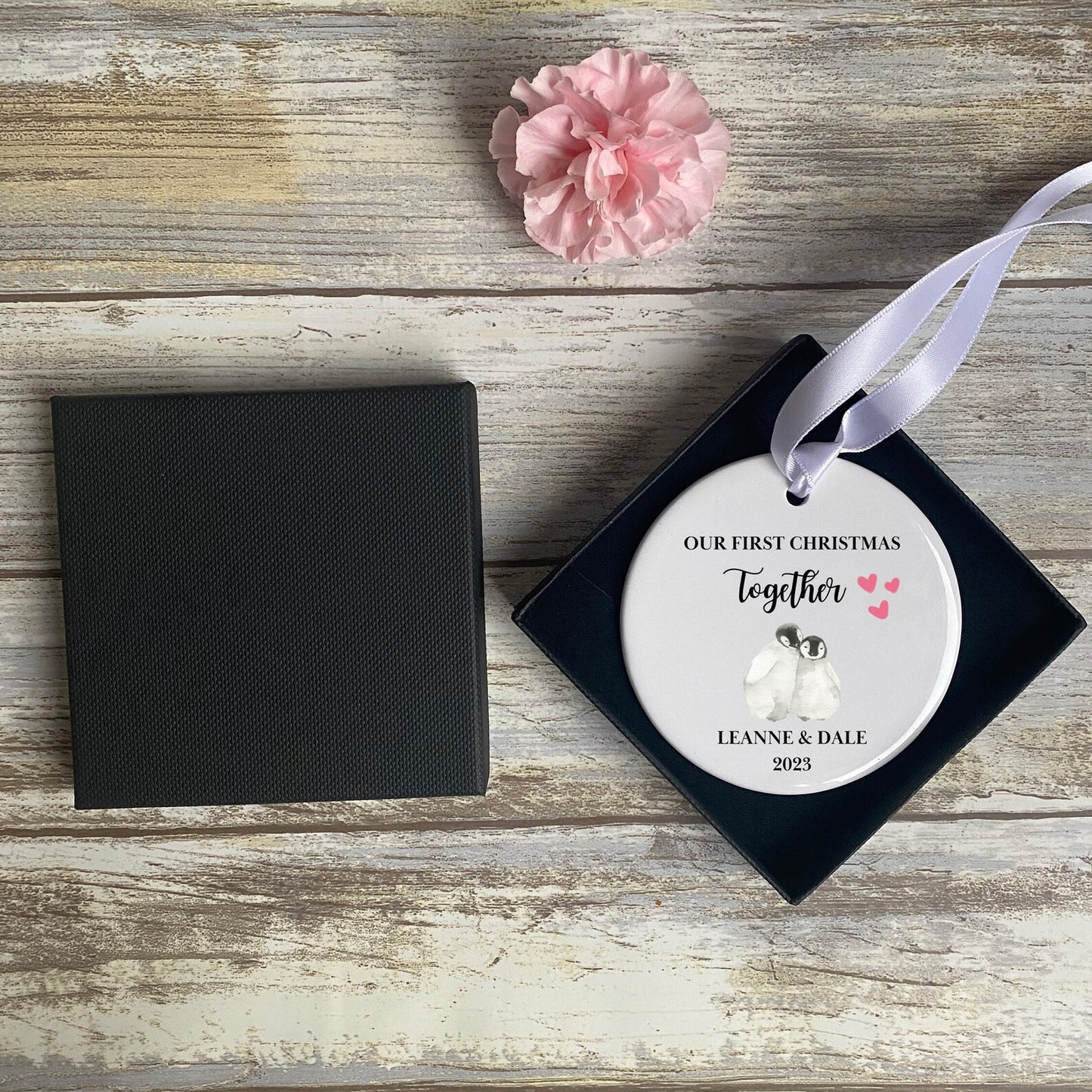 Flat lay of personalised 'Our First Christmas Together' bauble featuring two penguins, pink hearts, couple's names and 2023 date – romantic keepsake ornament in gift box, perfect for newly engaged or married couples