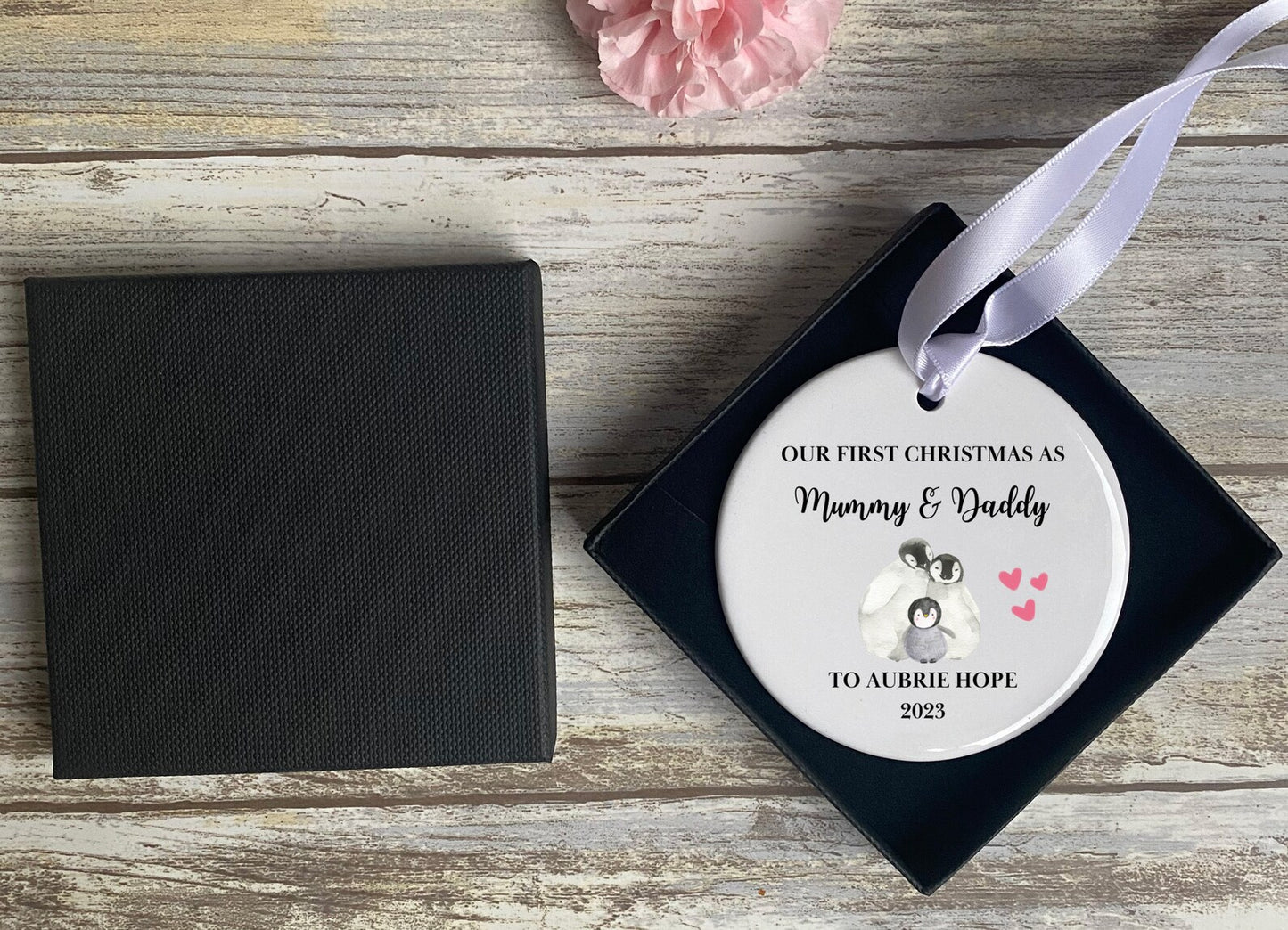 Personalised ceramic Christmas bauble in gift box, featuring 'Our First Christmas as Mummy & Daddy' text, baby name 'Aubrie Hope', penguin family illustration with pink hearts, and 2023 date – thoughtful new parents keepsake decoration