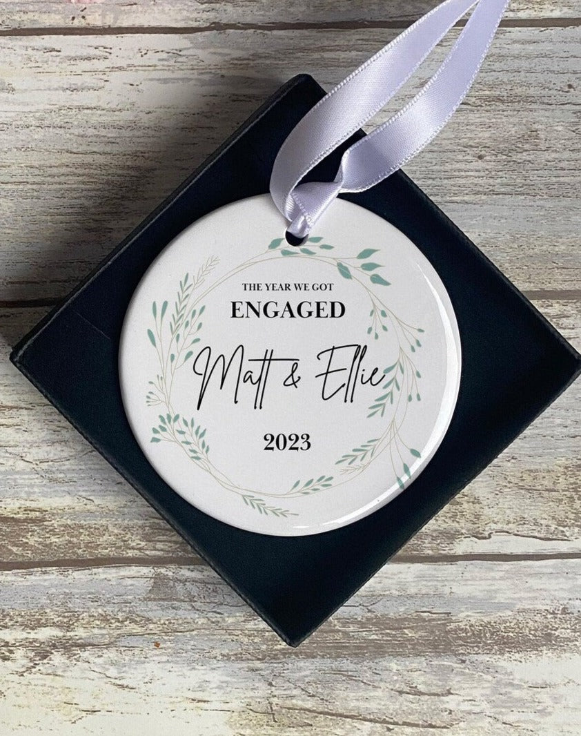 Personalised ceramic engagement Christmas ornament in a gift box, featuring 'The Year We Got Engaged', names 'Matt & Ellie', and the year 2023 surrounded by a delicate green botanical wreath design – elegant keepsake decoration.