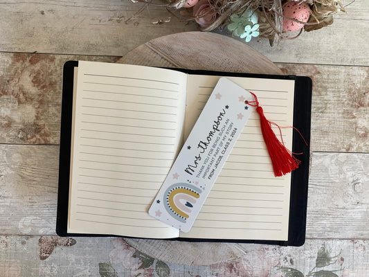 Open lined notebook with a personalized bookmark placed diagonally across the pages. The bookmark features the name 'Mrs Thompson' in cursive, a thank you message from a student, and a red tassel. The background includes a rustic wooden surface with soft floral and Easter egg decorations.
