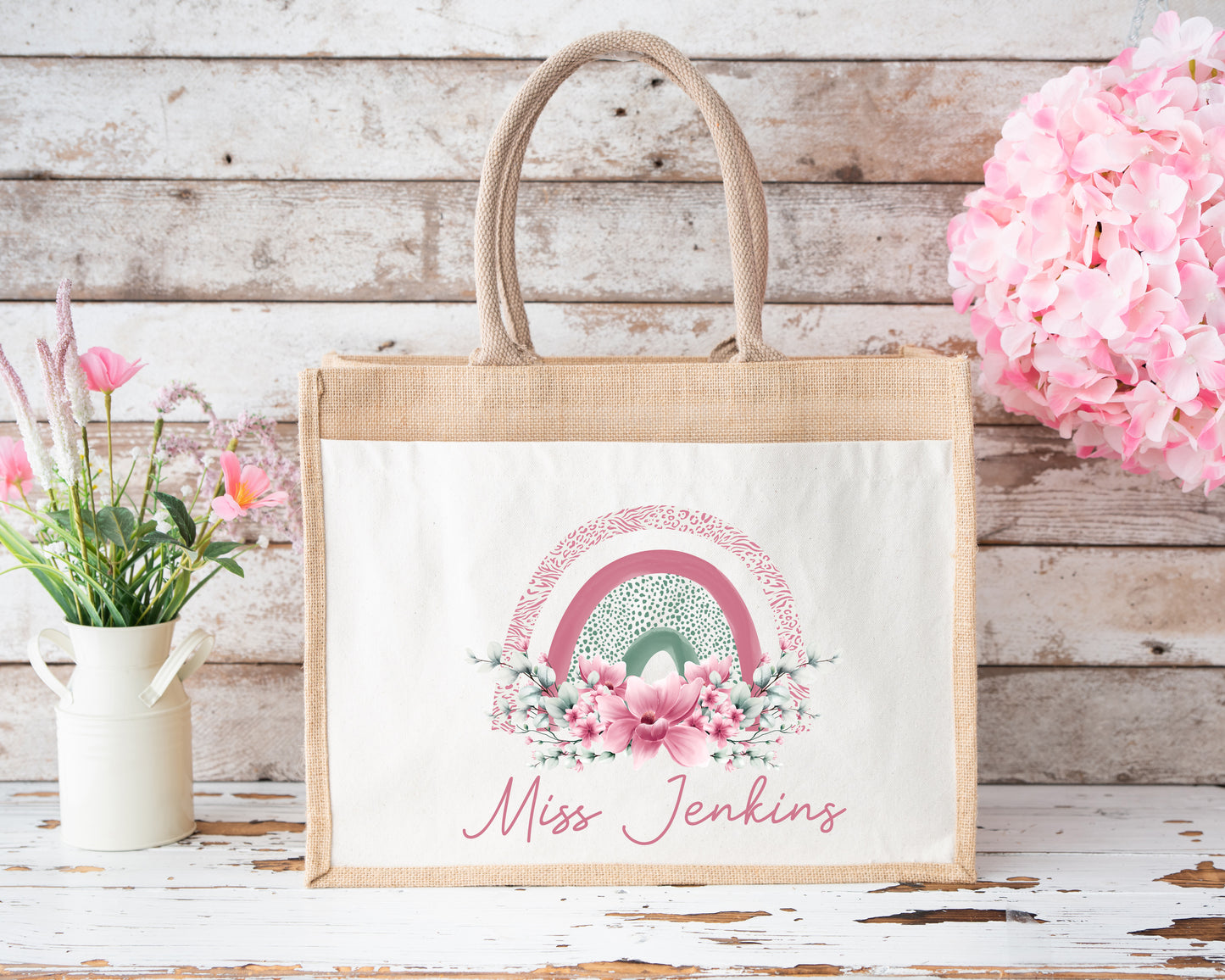 Rustic-style jute tote bag with āMiss Jenkinsā written in elegant pink script. The bag features a pastel rainbow and floral design in shades of pink and green. It is set on a distressed white wooden background with pink and white flowers arranged decoratively around it.