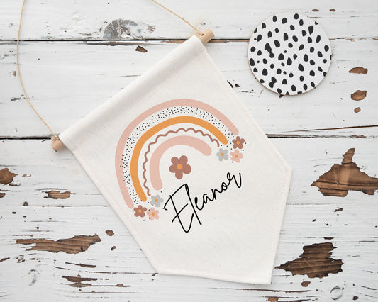 A soft linen banner hanging on a rustic wooden surface, featuring the name “Eleanor” in elegant black script below a playful rainbow illustration in warm tones of pink, mustard, and terracotta. The rainbow is accented with floral elements and dotted patterns, giving it a whimsical, boho feel. A monochrome spotted coaster lies nearby.