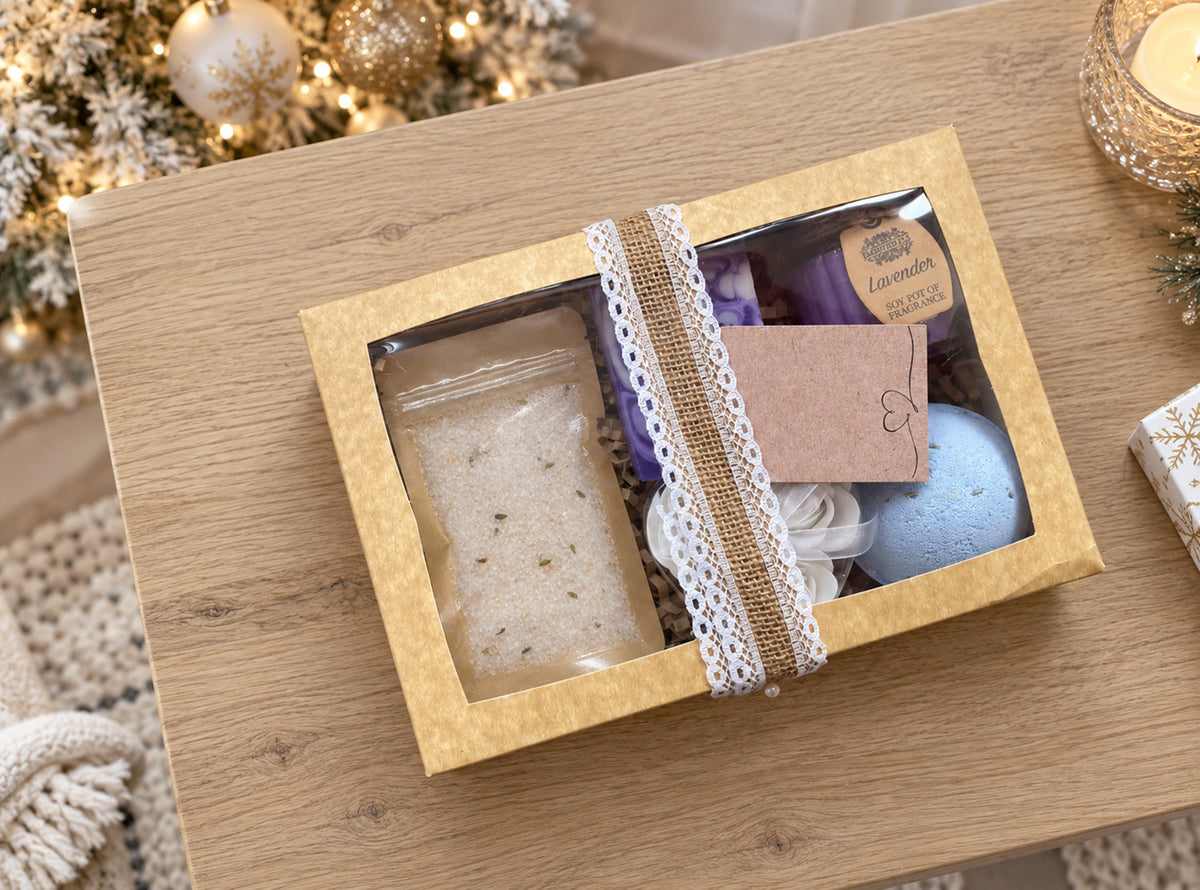 A gift box of bath and body items displayed on a wooden table, featuring a clear window showing a beige bath salt packet, a lavender soap bar labeled Lavender, a blue bath bomb, and decorative burlap and lace ribbon accents.