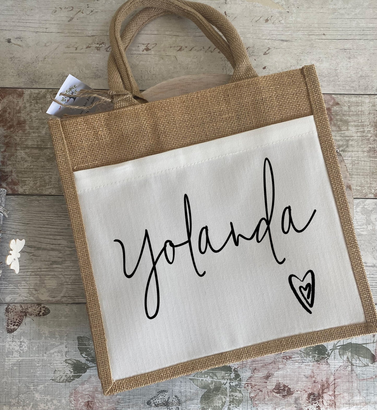 A rustic jute bag with white canvas front panel featuring the name "Yolanda" in stylish black cursive font. A heart graphic appears at the bottom right. The bag is placed on a distressed floral wood background with butterflies and roses.