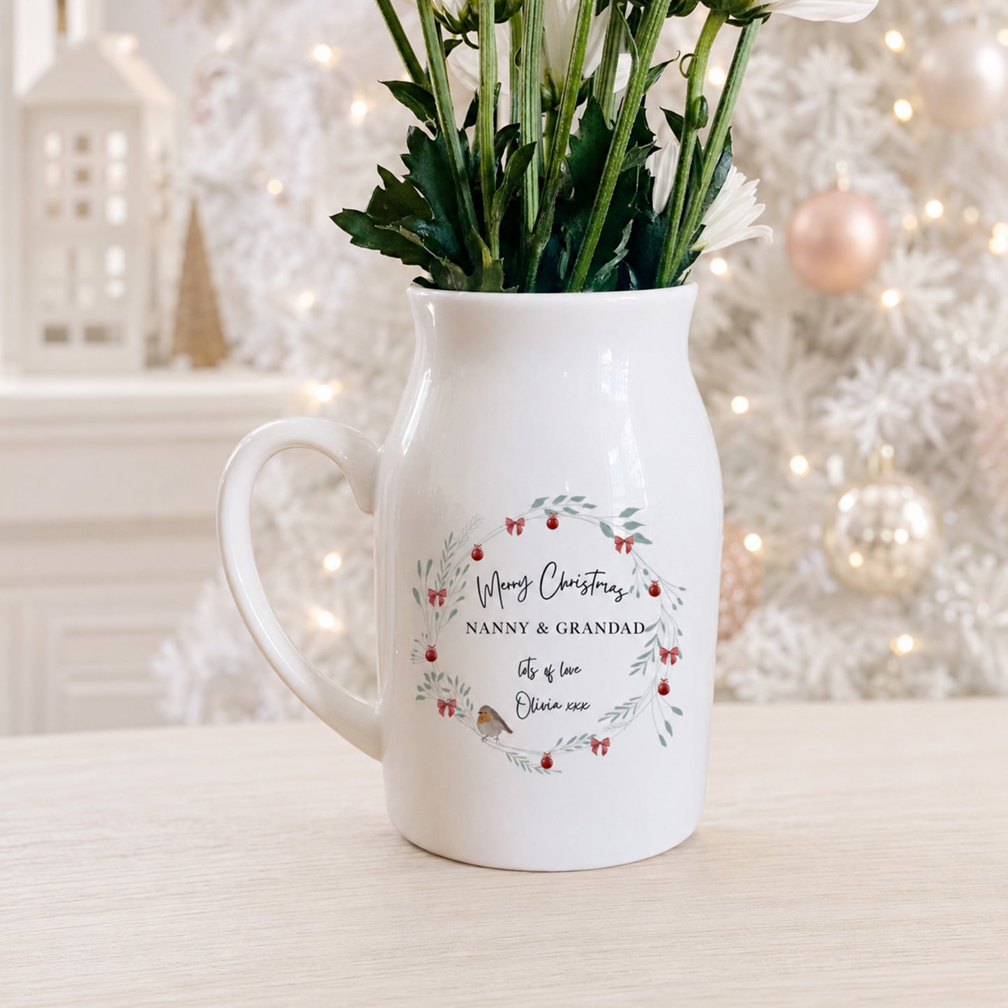 A white ceramic Christmas pitcher vase with a handle, filled with green stems and white flowers, set on a light wooden table. The front of the vase features a personalized wreath design with red berries, small birds, and the text 'Merry Christmas NANNY & GRANDAD, life is love Eleanor'.