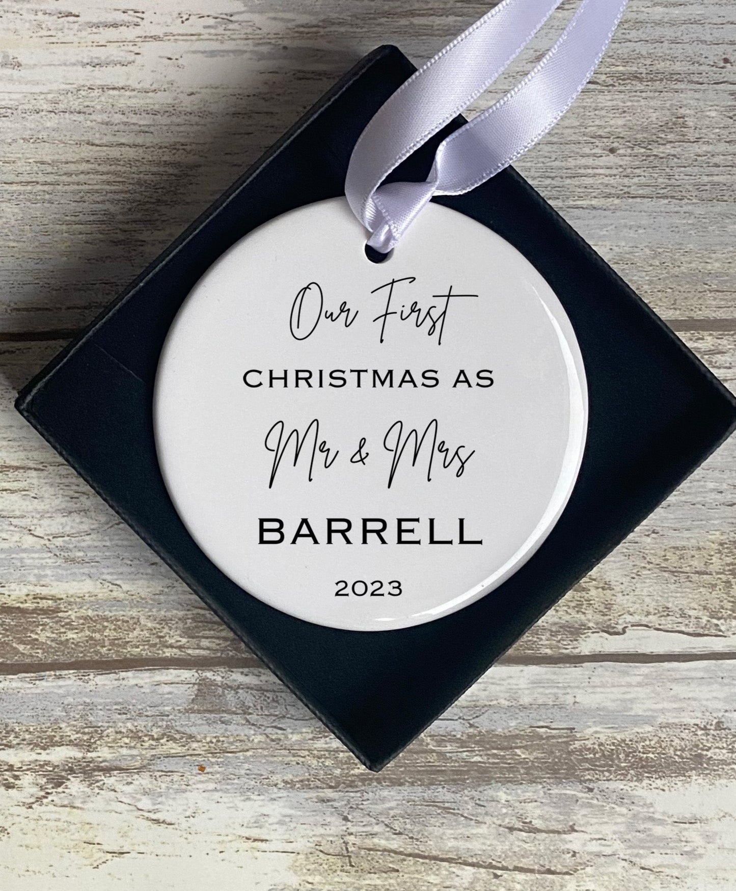 Ceramic Christmas ornament in a navy blue gift box with a white satin ribbon, displaying the text “Our First Christmas As Mr & Mrs Barrell 2023” in a mix of elegant script and bold serif fonts. The ornament rests on a rustic wooden background.