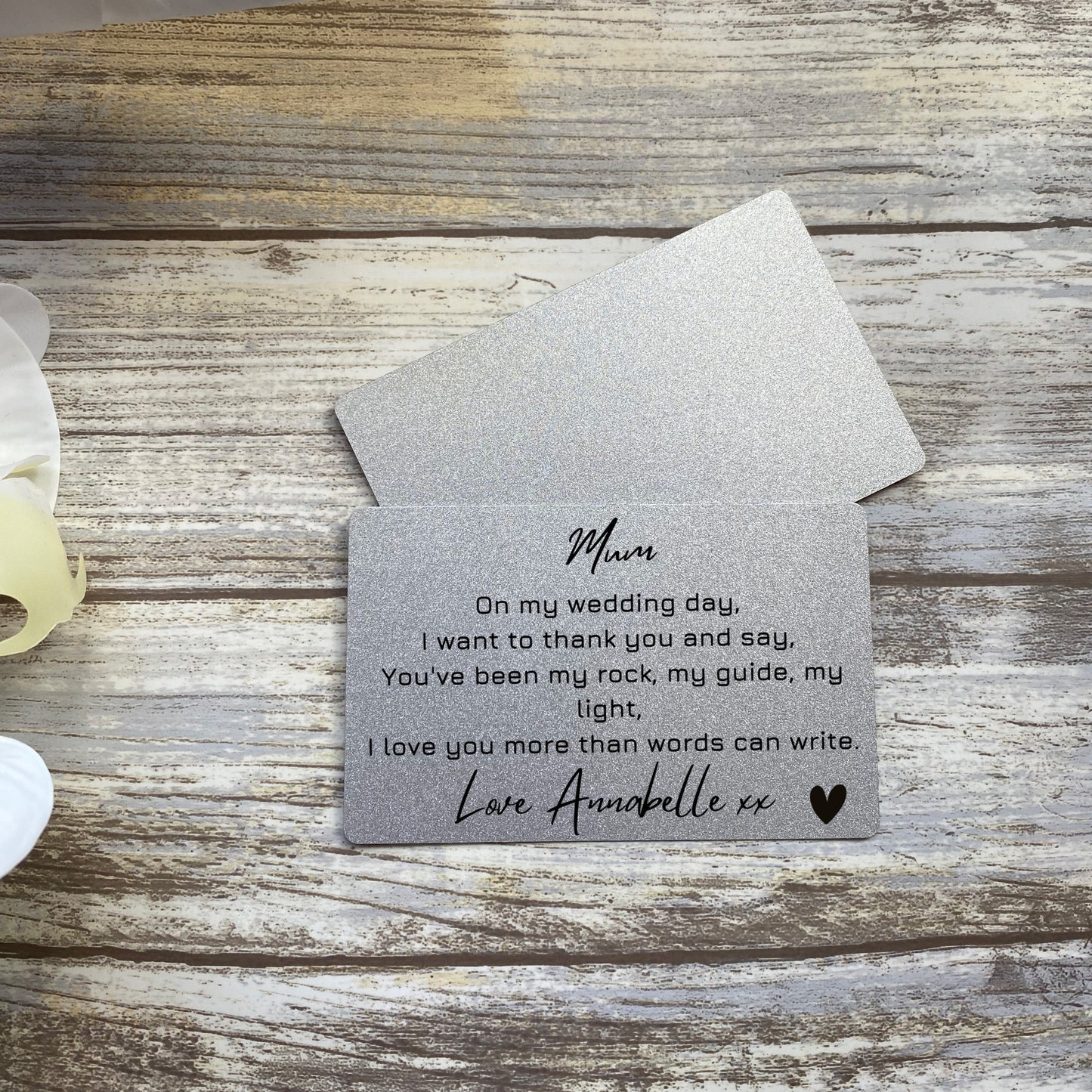 A silver metallic wallet-sized card placed on a rustic wooden surface. The card contains a heartfelt message from a daughter to her mother, expressing love and gratitude for guidance and support. The message is printed in black text, signed with a name and heart symbol. An envelope rests partially visible behind the card.